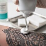 Zbliżenie na dłoń wyciskającą biały krem z tubki na świeży, czarny tatuaż.