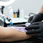 Skupienie na dłoniach tatuatora ubranego w czarne rękawiczki, który precyzyjnie przykleja fioletową kalkę ze wzorem na zdezynfekowane przedramię klienta