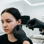 Zbliżenie na dłonie w czarnych rękawiczkach nitrylowych, które precyzyjnie chwytają tytanowy kolczyk (labret) na płatku ucha.