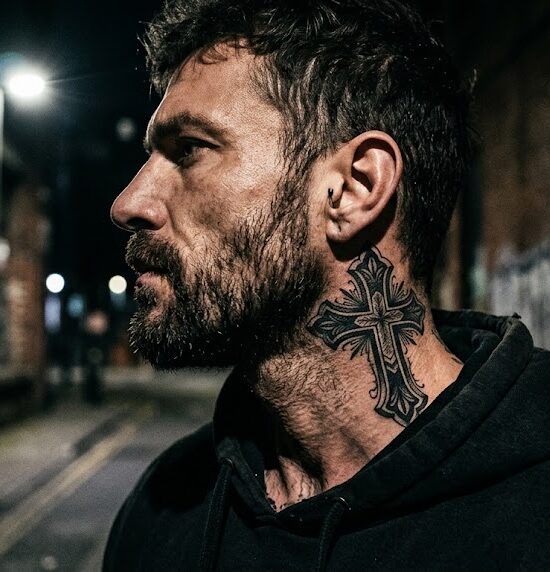 Na bocznej części szyi widnieje mocny, czarny tatuaż krzyża w stylu Blackwork