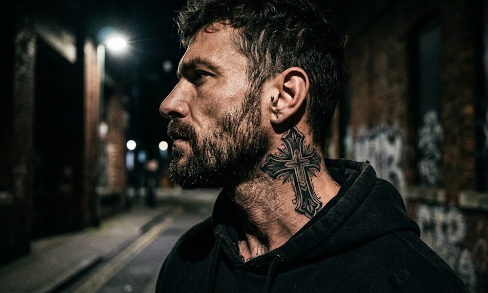 Na bocznej części szyi widnieje mocny, czarny tatuaż krzyża w stylu Blackwork